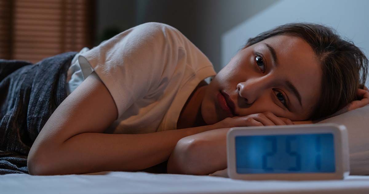Woman laying in bed with alarm clock in front of her.