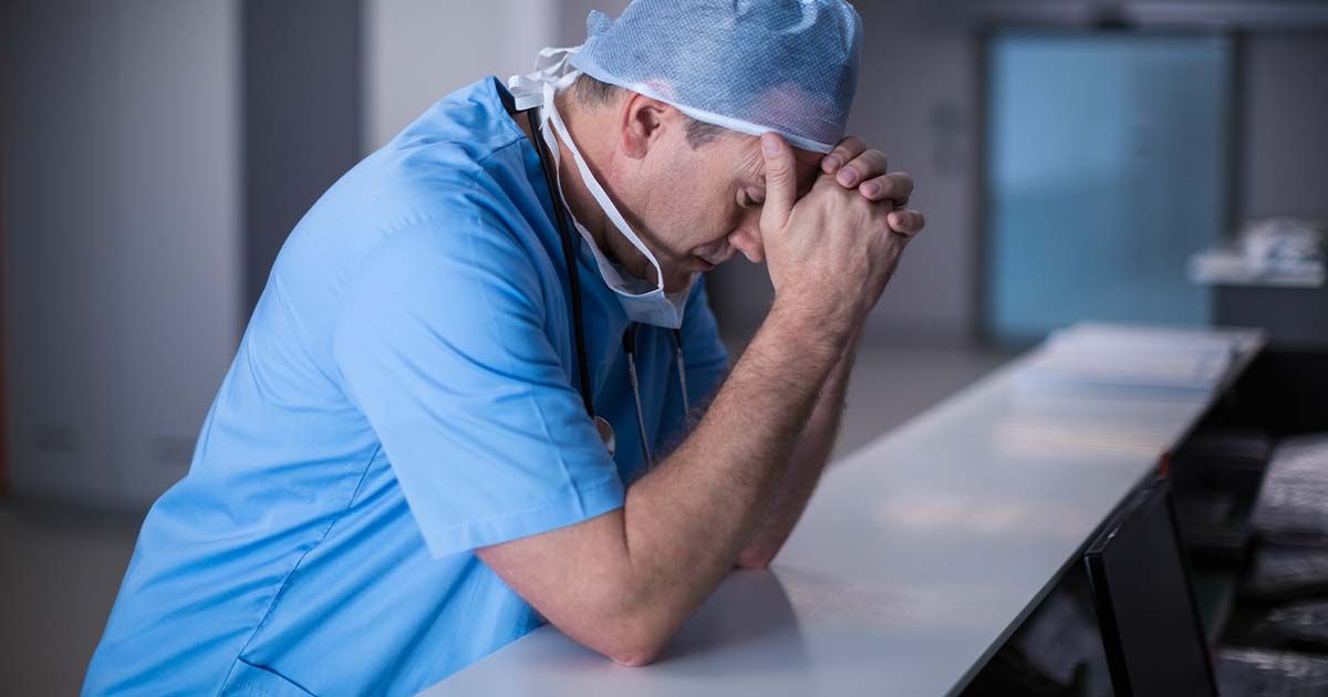 Surgeon in hospital at front desk with hands on forehead looking tired.