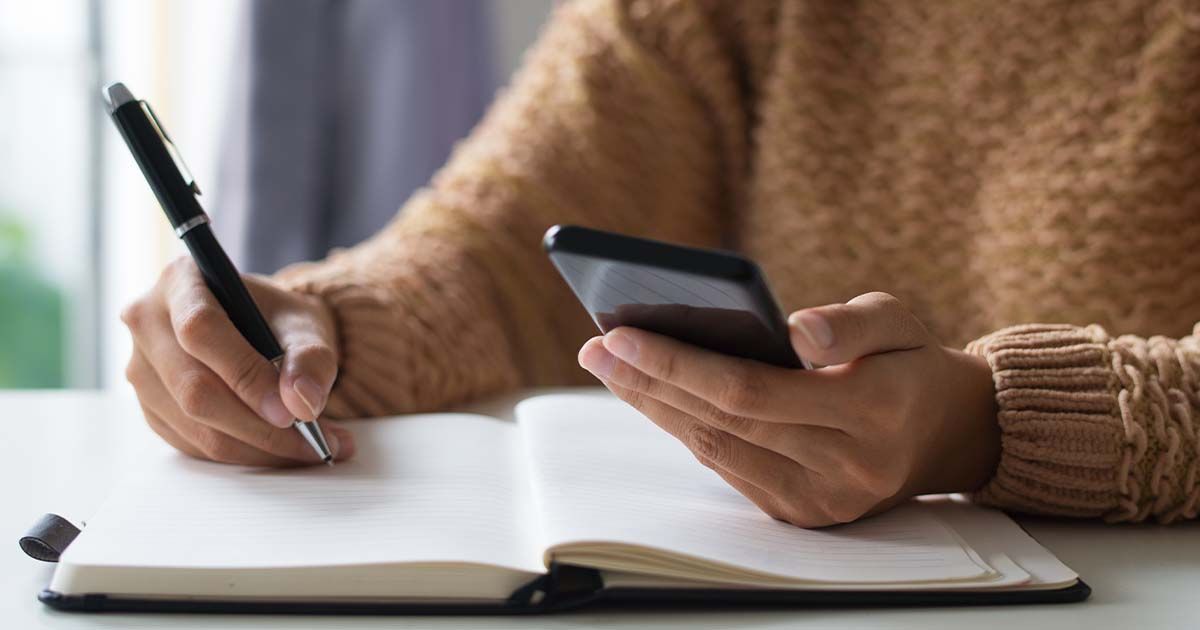 Woman looking at phone and writing in journal/schedule.